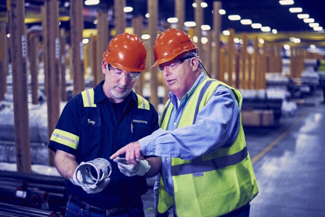 Marmon Keystone Two men wearing orange hard hats and safety glasses stand in a factory. One holds a metal tube as the other, in a safety vest, points at it, discussing or inspecting the item as part of the supplychain in a metals distributor setting.