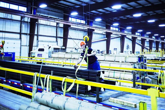 Marmon Keystone A worker in a hard hat and safety vest operates machinery to move long metal bars inside a large industrial warehouse, supporting the metals supply chain under bright overhead lights and high ceilings.