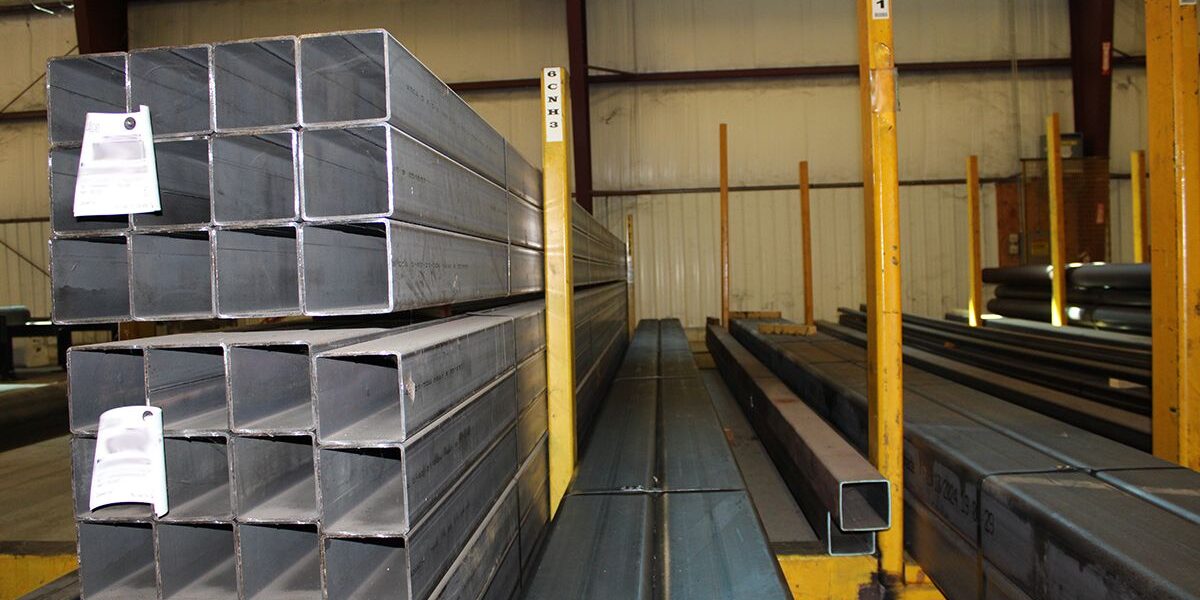 Marmon Keystone Stacks of rectangular steel tubing are arranged on racks inside a warehouse. A Marmon/Keystone sign is displayed at the top, highlighting their role as a leading metals distributor. The words "IN STOCK HSS" appear at the bottom of the image.