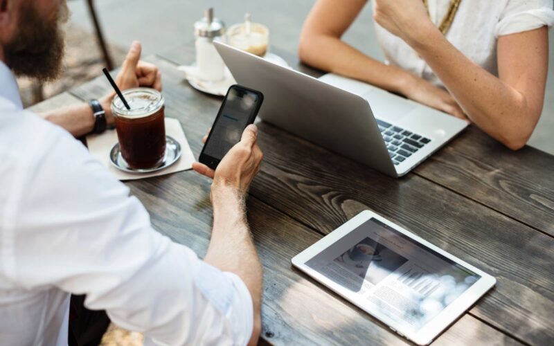 Marmon Keystone Two people sit at a wooden table outdoors with a laptop, tablet, smartphone, and iced coffee. One person types on the laptop while the other holds a phone. The tablet displays a metals supplychain distributor webpage.