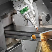Marmon Keystone A close-up view of a laser cutting machine cutting a circular hole into a metal tube, with bright sparks flying from the cut area. The machinery and metal surface are in sharp focus, showcasing precision in metals supplychain operations.