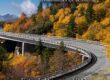 Marmon Keystone A curved highway bridge winds through a forest of autumn trees with vibrant yellow and orange foliage. Text promotes Marmon/Keystone aluminum pipe and tubing, stating Extruded Structural B429 is in stock and ready to ship.
