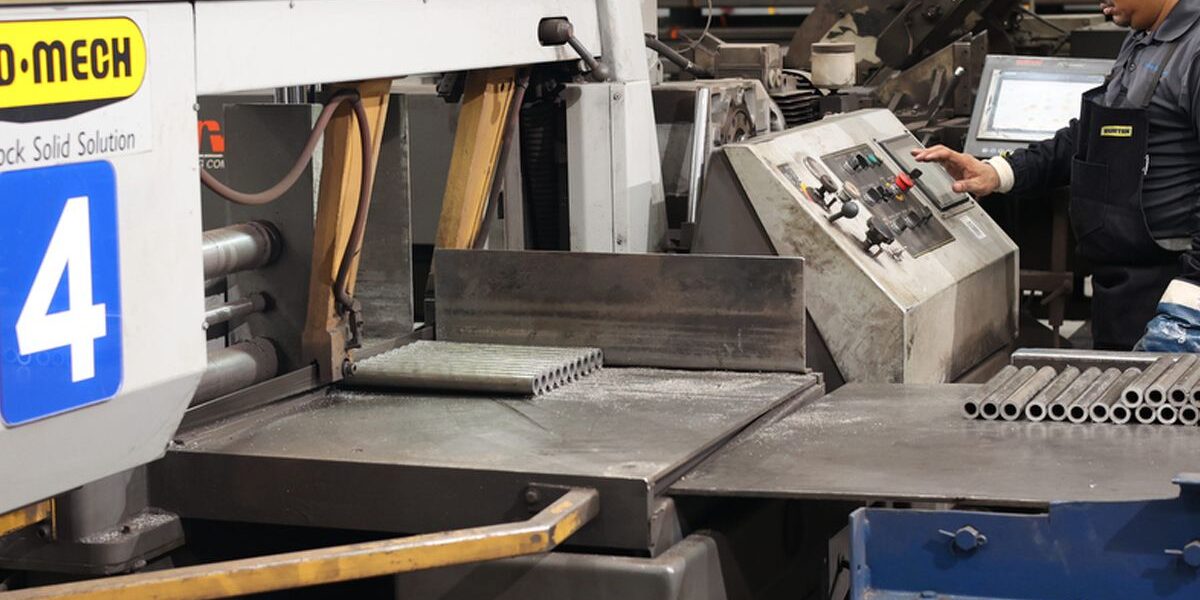 Marmon Keystone A worker operates industrial machinery in a factory setting by Marmon/Keystone. Metal tubes and equipment are visible, with signs in English and Spanish. Text on the image reads: "Marmon/Keystone delivers! en mexico.